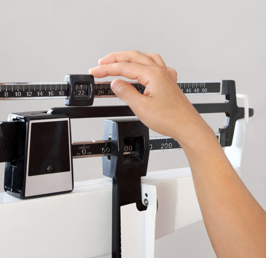 A person’s hand adjusts the sliding weight on a mechanical physician’s scale, commonly used for measuring body weight, against a plain light background.