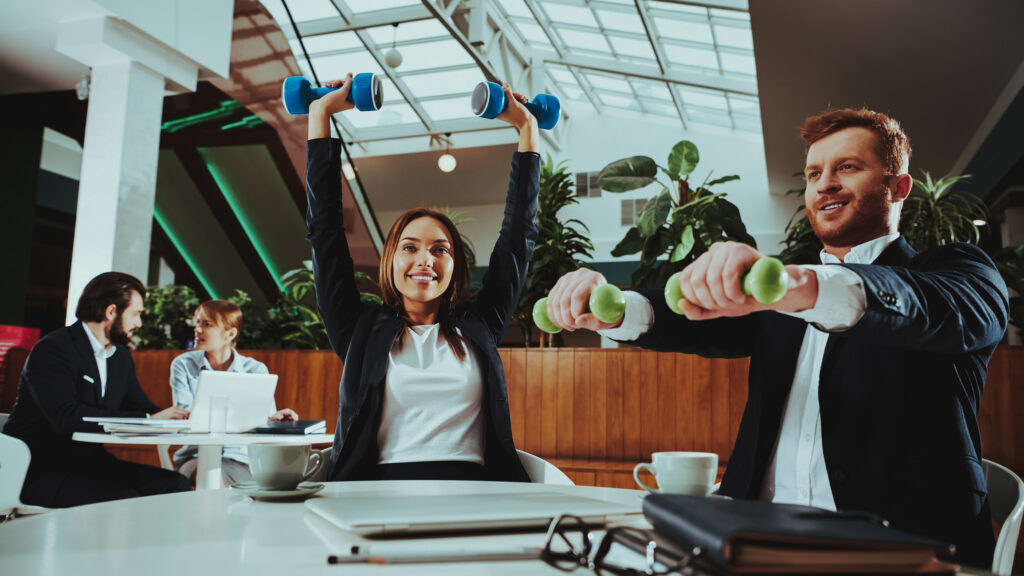 Two businesspeople in suits exercise with small dumbbells at a table in a modern office, smiling and surrounded by laptops, coffee cups, and paperwork. Other colleagues work in the background.