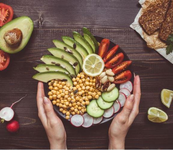 A person holds a bowl filled with sliced avocado, tomatoes, cucumber, radishes, chickpeas, nuts, and a lemon slice, with whole avocado, crispbread, and radishes on a dark wooden table.