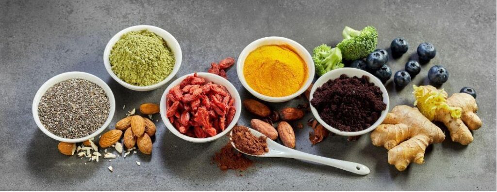 A variety of superfoods arranged on a gray surface, including bowls of chia seeds, green powder, goji berries, turmeric, acai powder, whole almonds, cacao beans, a spoon of powder, fresh broccoli, blueberries, and ginger root.