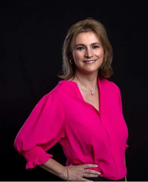 A woman with brown hair, wearing a bright pink blouse and a necklace, stands smiling with one hand on her hip against a black background.