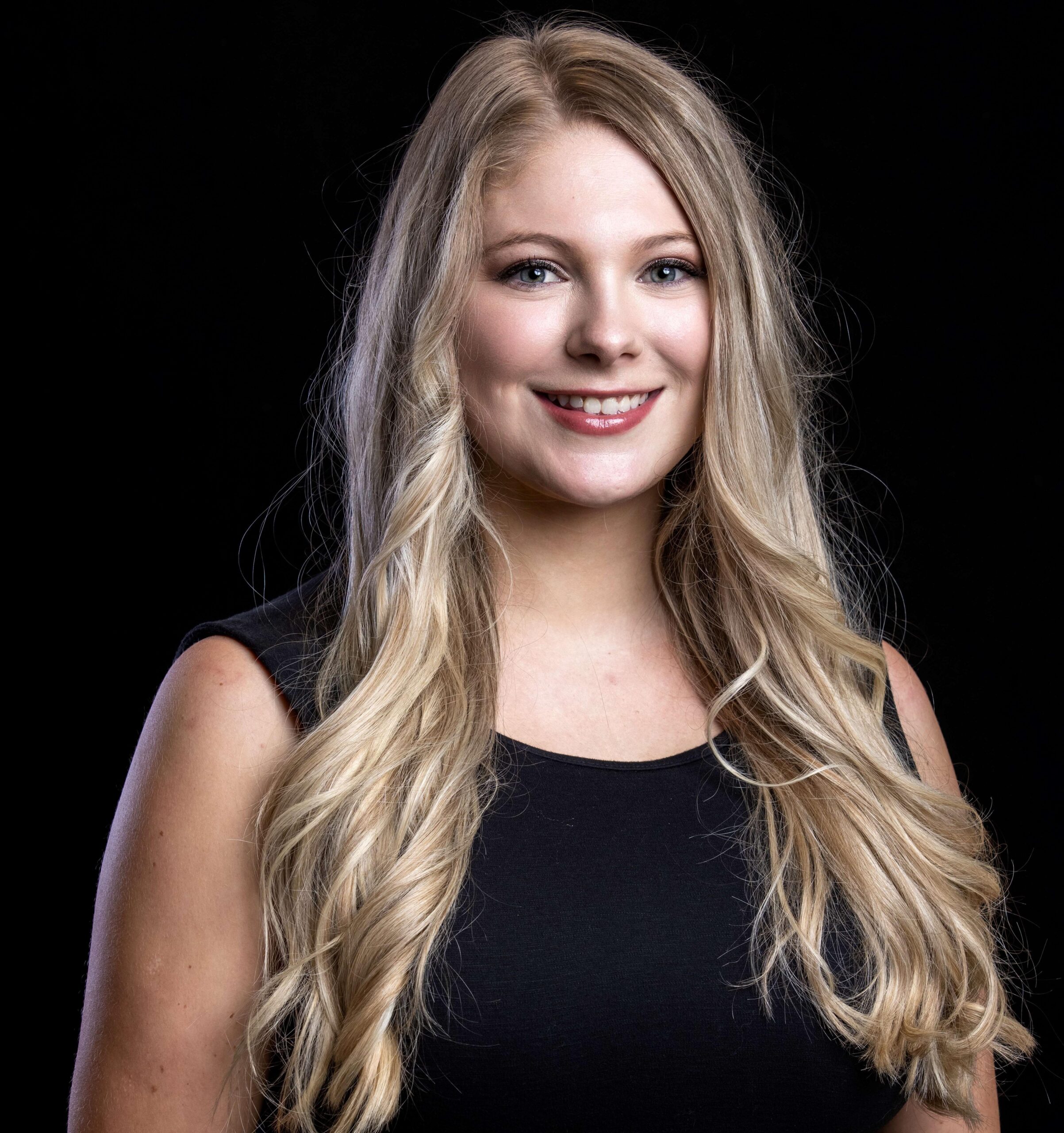 A young woman with long, wavy blonde hair smiles while wearing a sleeveless black top, standing against a solid black background.