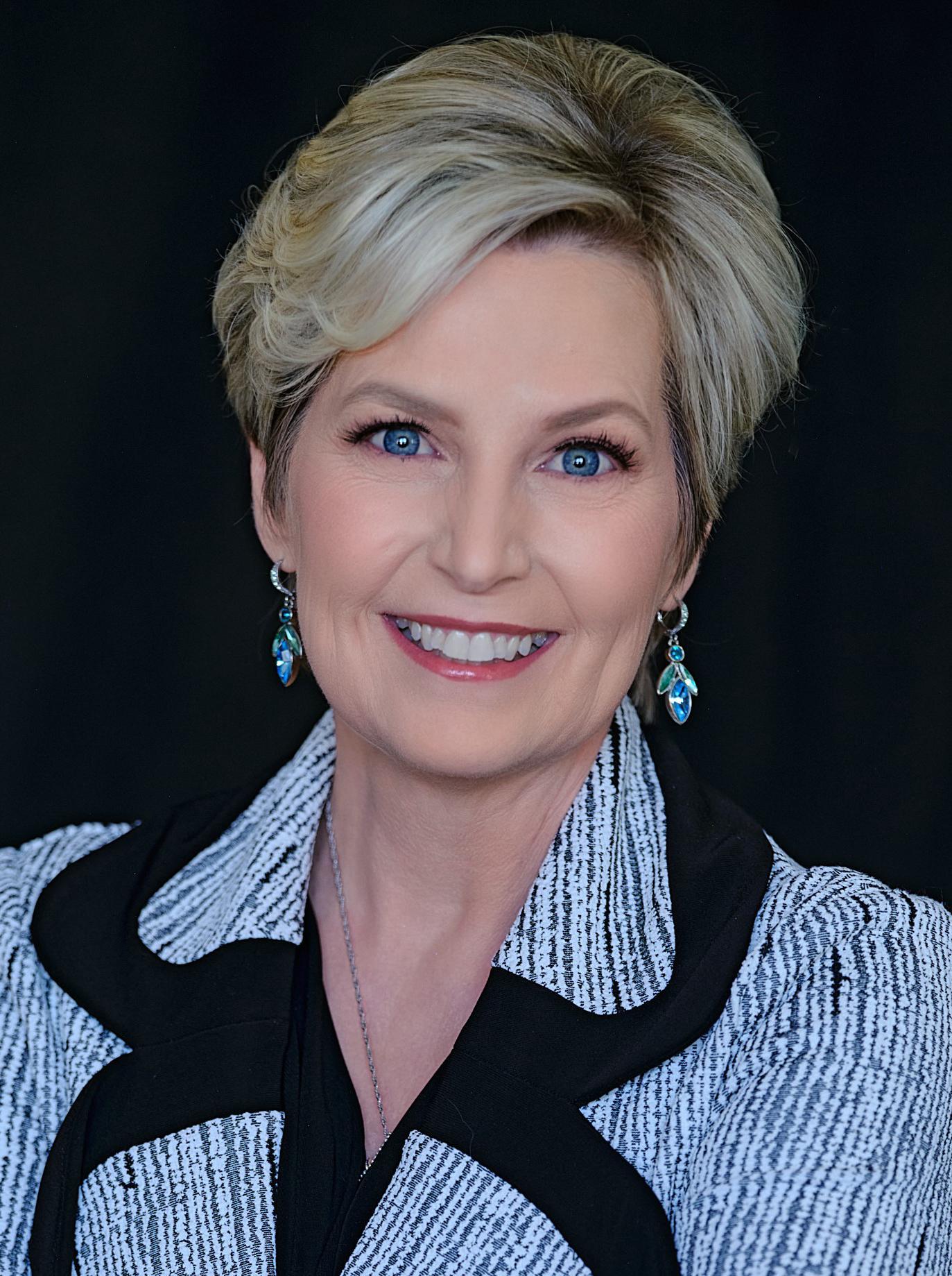 A woman with short, blond hair and blue eyes smiles at the camera. She is wearing turquoise earrings and a black and white textured jacket, posed against a dark background.