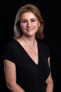 A woman with light brown hair wearing a black dress, gold hoop earrings, and a pearl necklace smiles while posing against a solid black background.