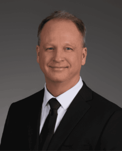 A middle-aged man with short light brown hair wearing a black suit, white shirt, and black tie, smiling at the camera against a plain dark background.