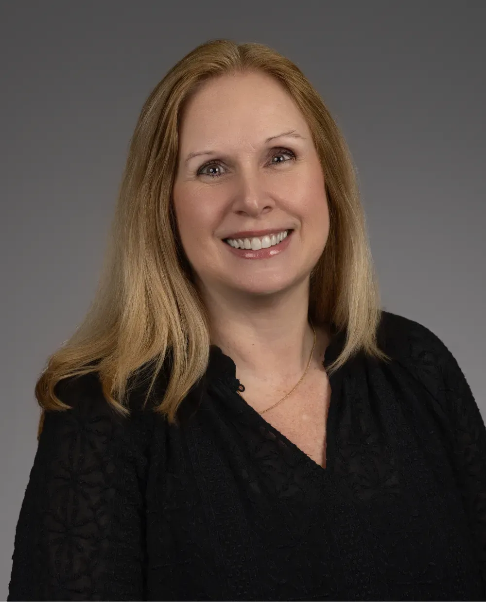 A woman with long blonde hair, wearing a black textured blouse and a gold necklace, smiles at the camera against a plain gray background.