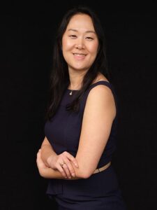 A woman with long dark hair, wearing a sleeveless navy dress and a thin beige belt, stands against a black background, smiling with her arms crossed.