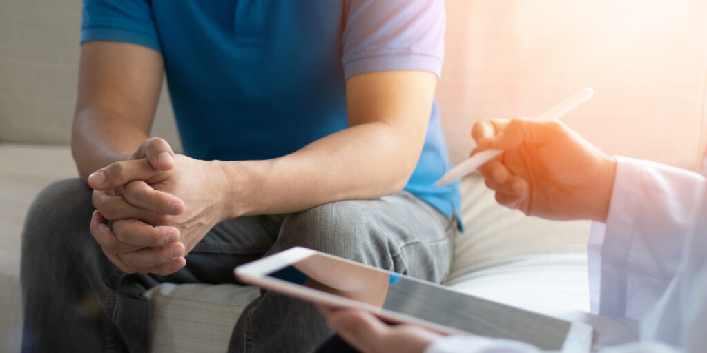 A person in a blue shirt sits with hands clasped, facing another person holding a digital tablet and stylus, suggesting a consultation or counseling session.