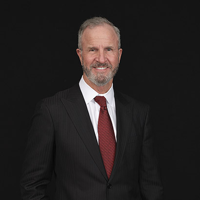 A middle-aged man with gray hair and a beard, wearing a black pinstripe suit, white shirt, and red tie, stands and smiles against a solid black background.