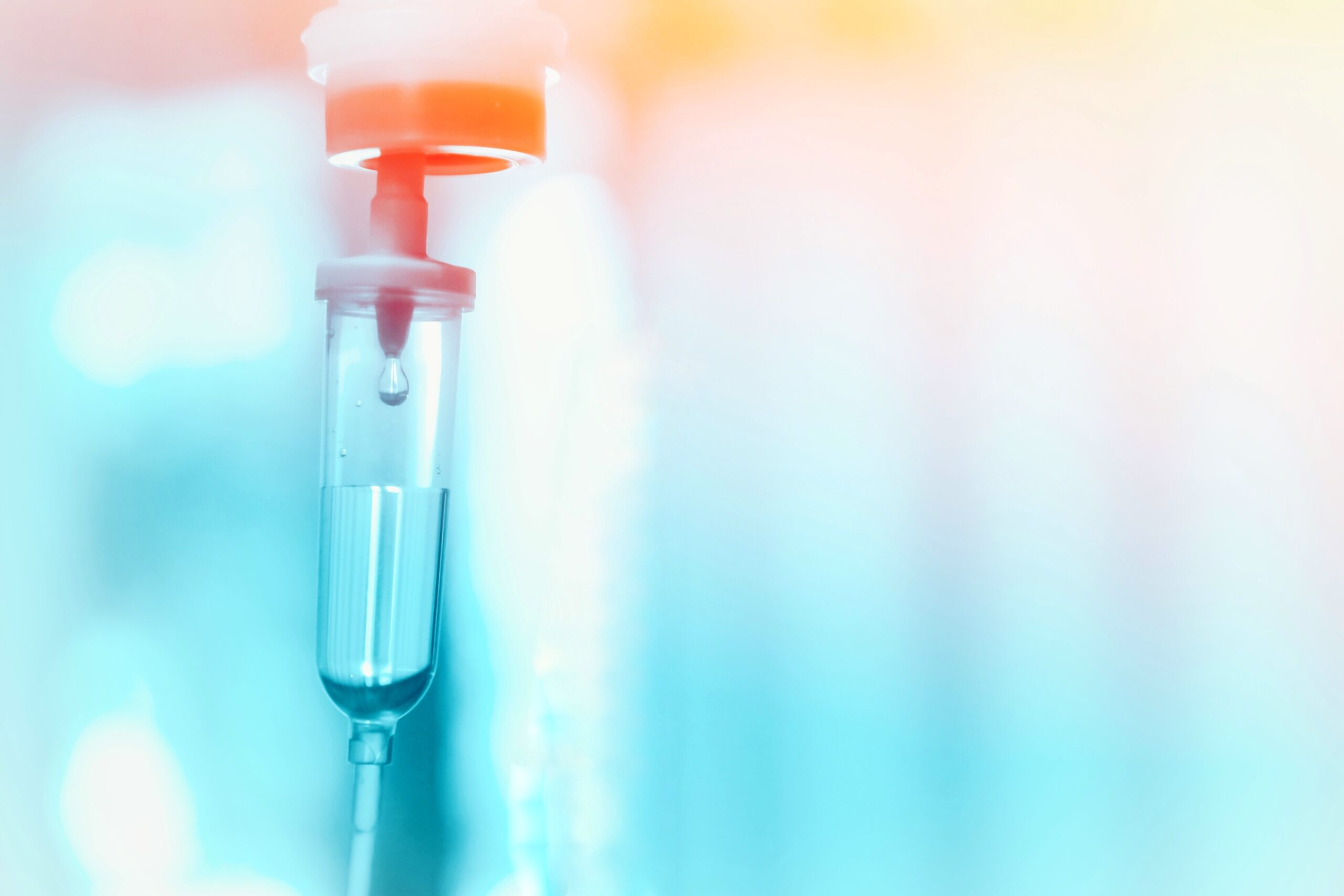 Close-up of an IV drip chamber with a droplet of fluid inside, against a blurred blue and orange background, suggesting a clinical or hospital setting.