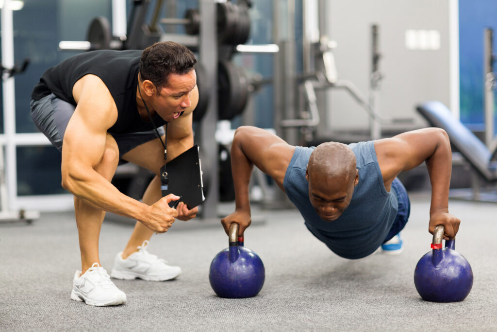 A man does push-ups gripping two kettlebells while a trainer kneels beside him, encouraging and guiding him in a gym setting with various exercise equipment in the background.