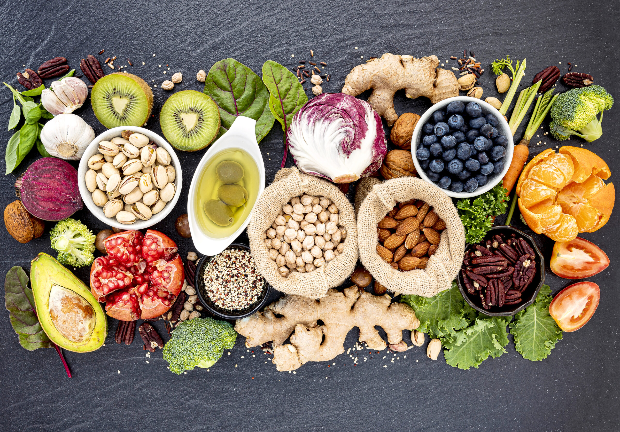 A colorful assortment of healthy foods including nuts, seeds, fruits, vegetables, leafy greens, and olive oil arranged on a dark surface. Items include blueberries, oranges, kiwi, avocado, chickpeas, tomatoes, broccoli, and garlic.