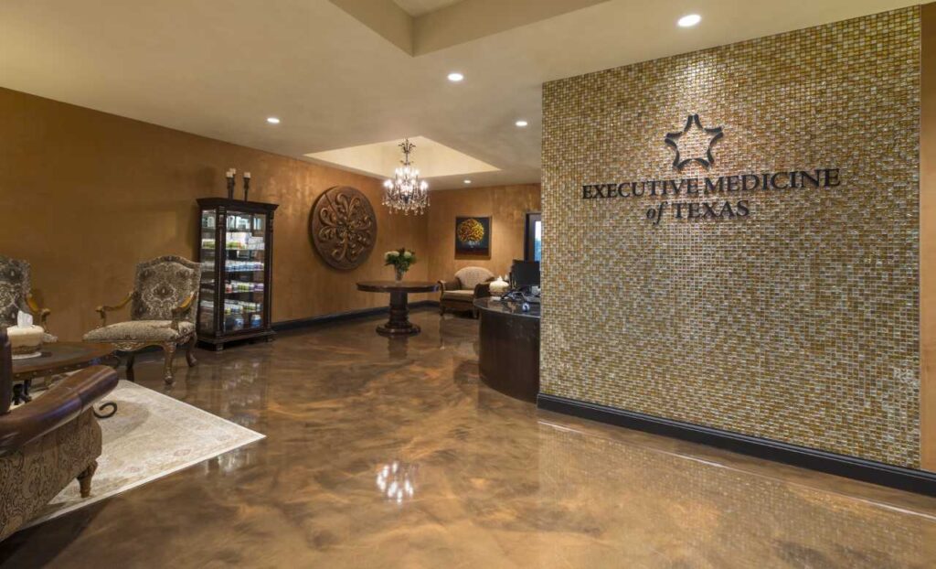 A luxurious medical office lobby with glossy brown floors, elegant chairs, a chandelier, wall art, and a mosaic-tiled wall displaying the logo and name Executive Medicine of Texas.