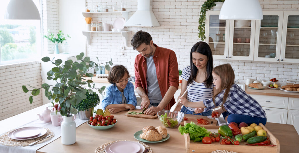 A family of four stands in a bright kitchen preparing a meal together, cutting vegetables and smiling. Fresh produce and bread are on the counter, with light streaming through large windows.