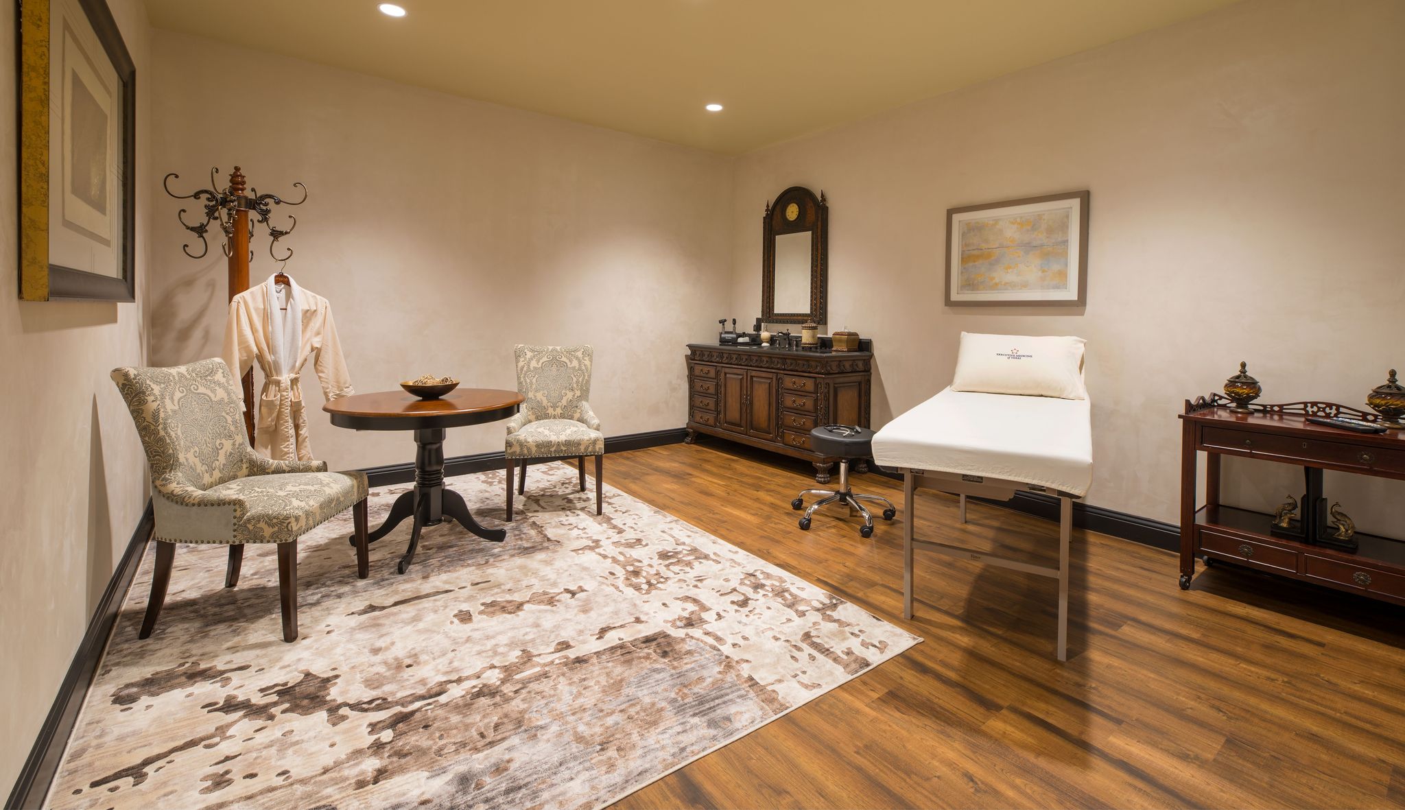 A spa treatment room with a massage table, two upholstered chairs and a small round table, a wooden dresser with a mirror, a coat rack holding a robe, and warm, soft lighting. The room has wooden floors and a patterned rug.
