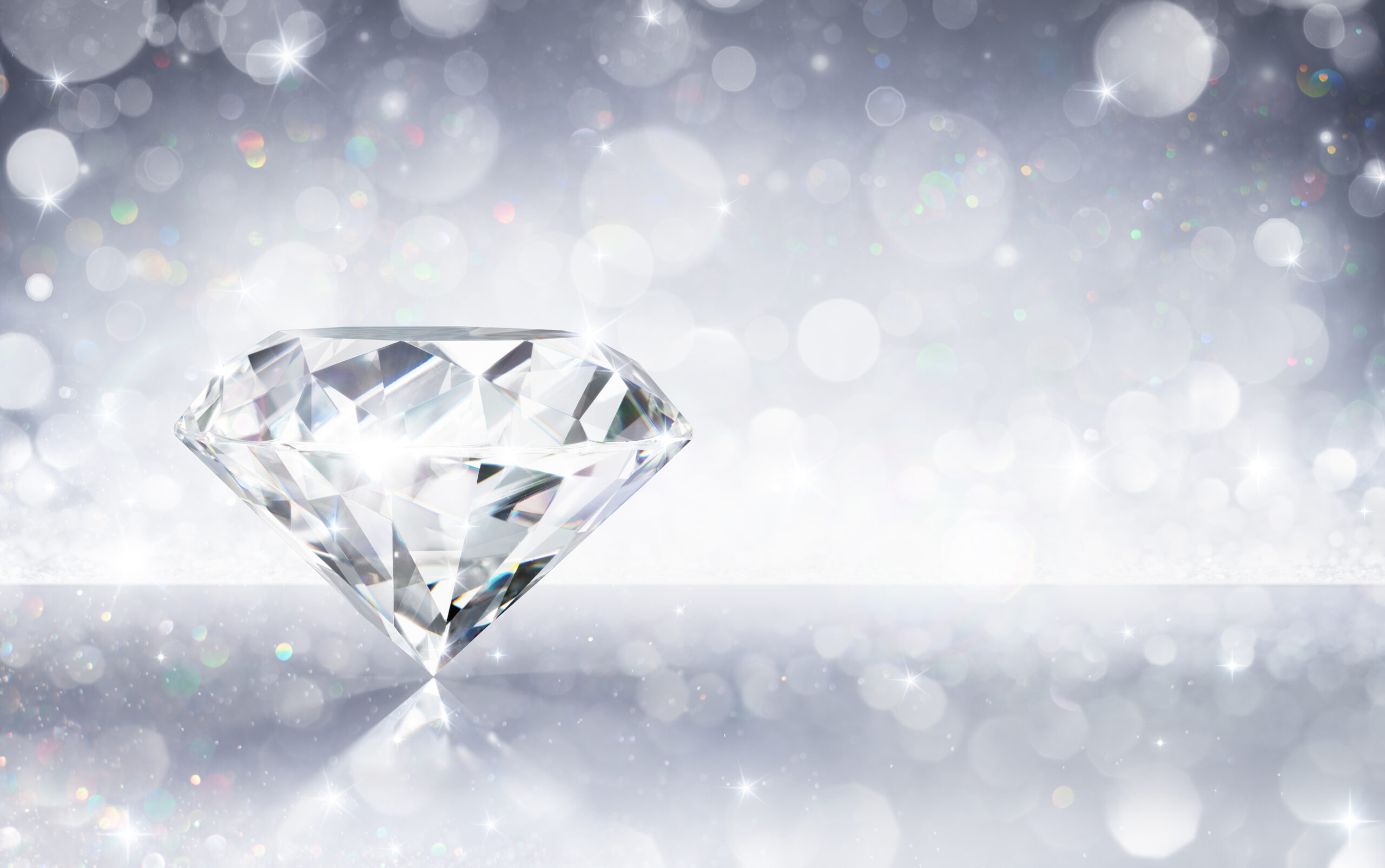 A sparkling diamond sits on a reflective surface with a soft, blurred background of white and silver bokeh lights, creating a shimmering and elegant atmosphere.