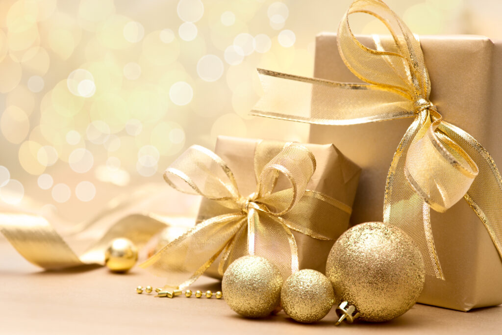 Three gold-wrapped gift boxes with gold ribbons are arranged beside three glittery gold Christmas ornaments and gold beads, all set against a softly blurred, golden festive background.