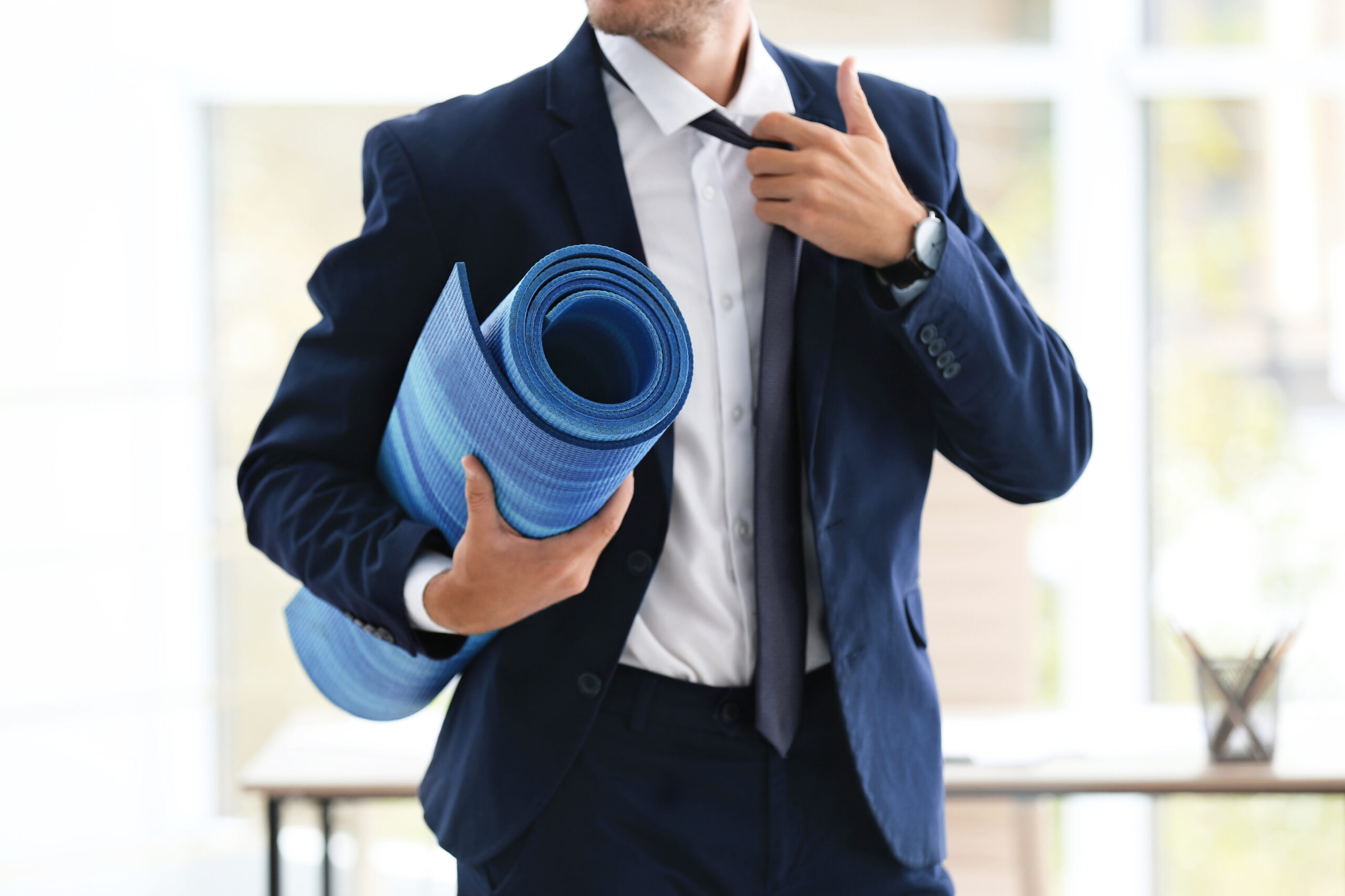 A man in a navy blue suit holds a rolled-up blue yoga mat under one arm while adjusting his tie, standing in a bright office space.