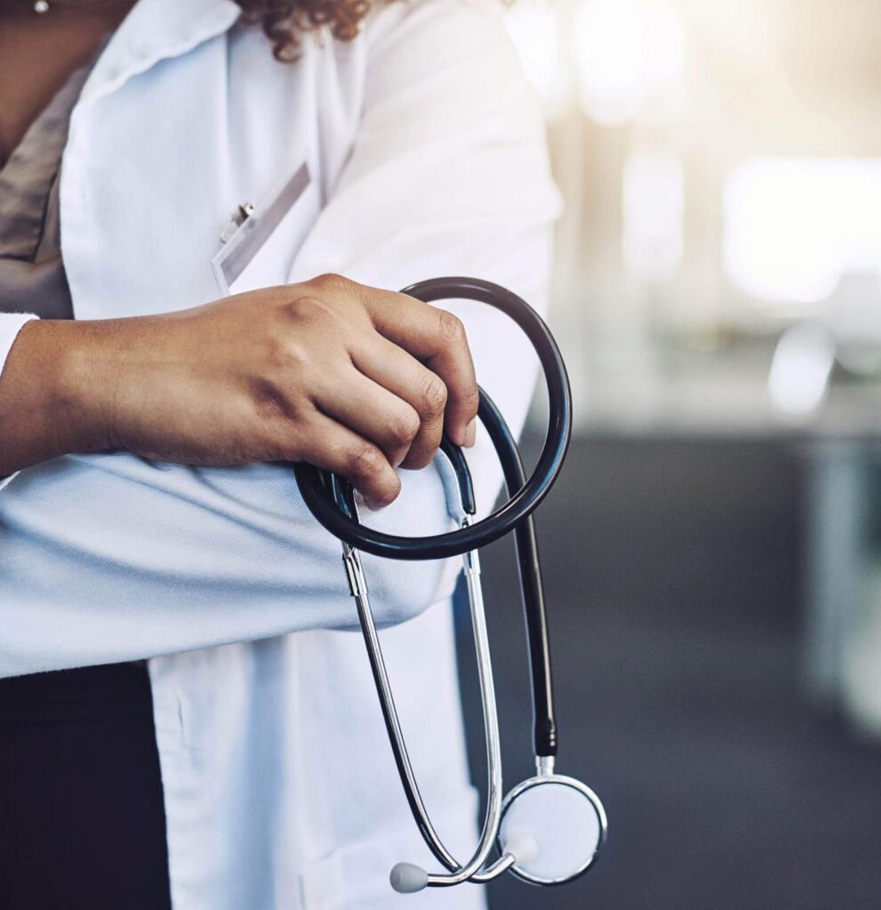 A close-up of a healthcare professional wearing a white coat and holding a stethoscope, with arms crossed and a blurred background.