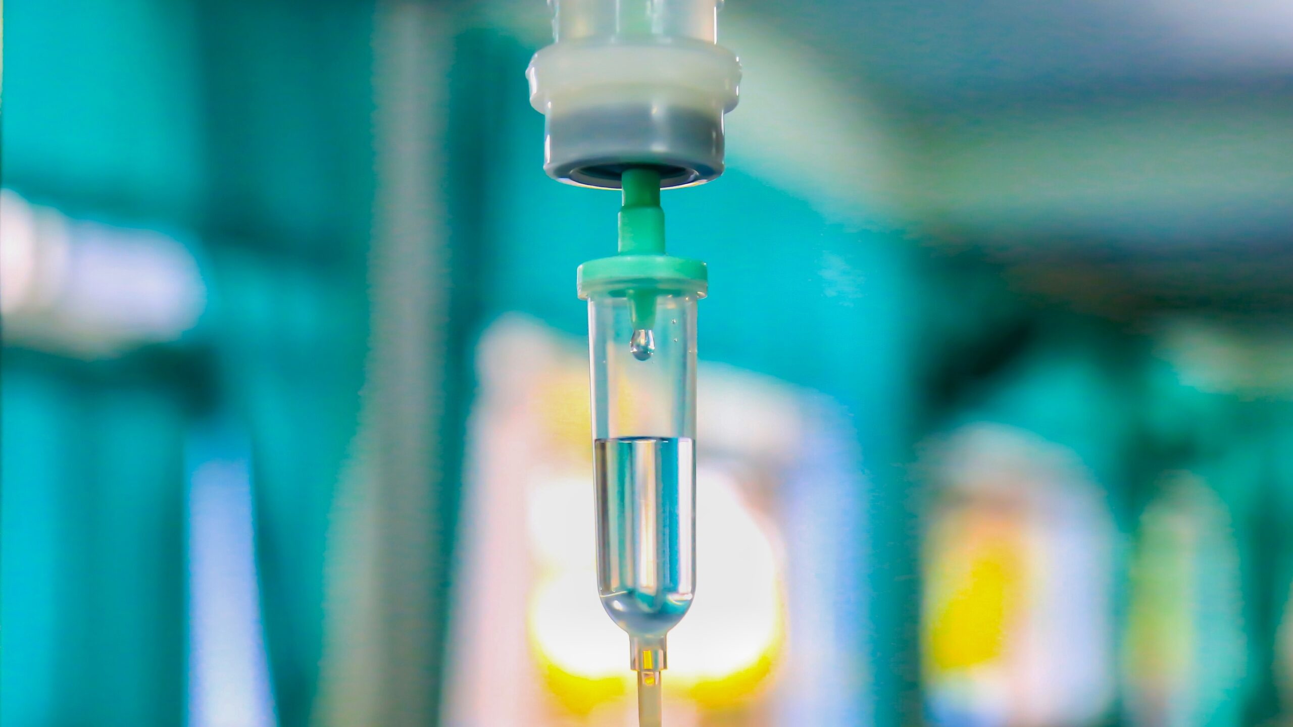 A close-up of an intravenous (IV) drip chamber filled with clear fluid, with a blurred hospital or medical setting in the background.