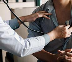 A doctor uses a stethoscope to listen to a patients chest, who is pulling aside their shirt for the examination.