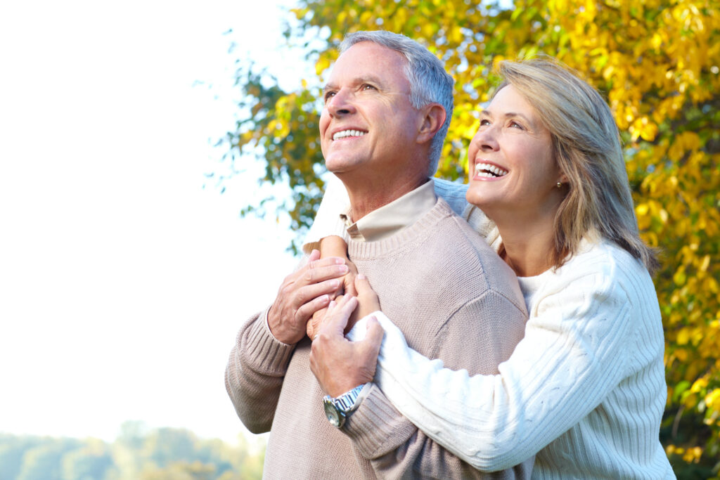 Smiling older couple outdoors, wearing sweaters, standing close together. The woman hugs the man from behind. Bright autumn trees with yellow leaves are in the background. Both look happy and content.
