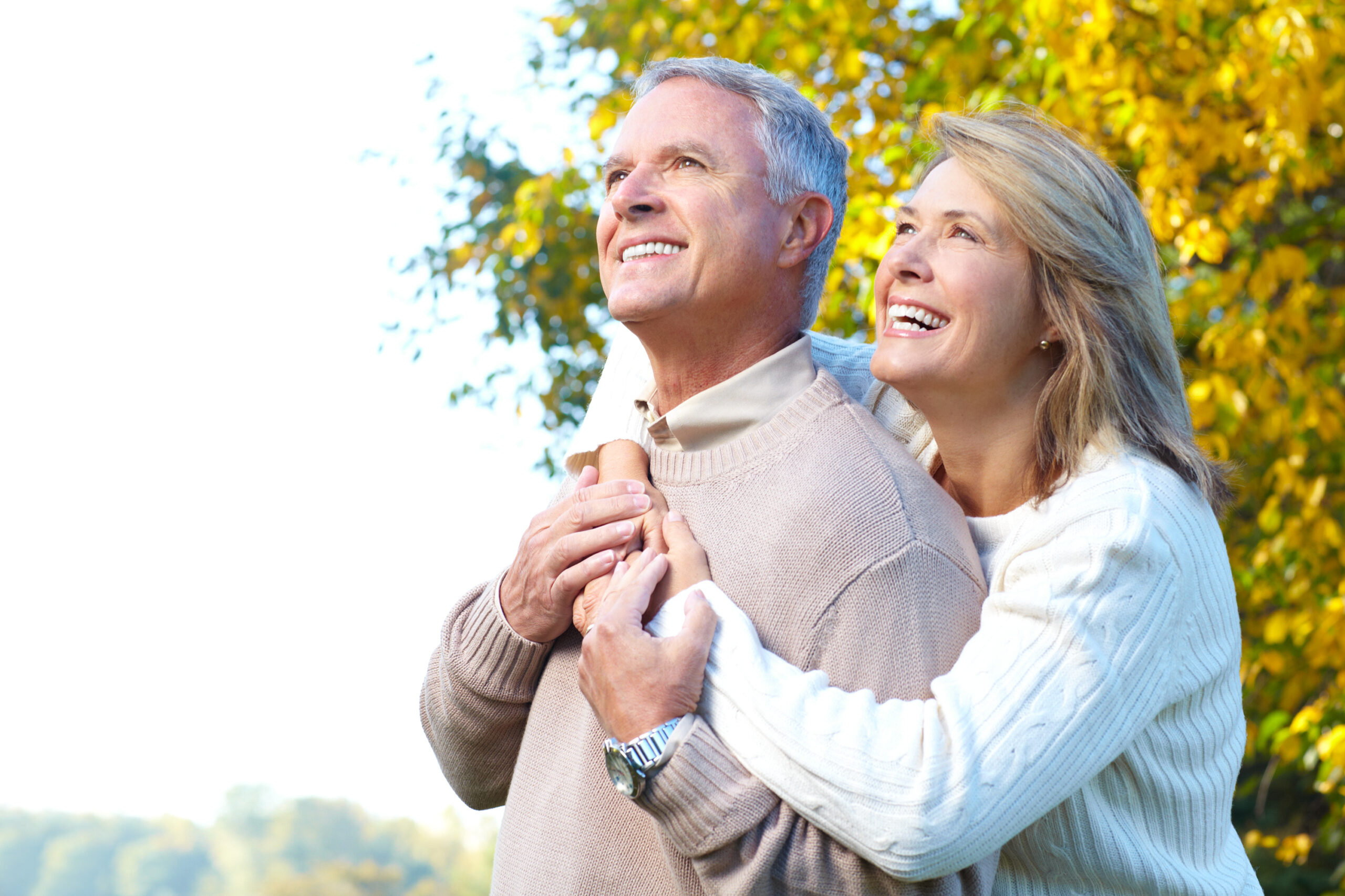 Smiling older couple outdoors, wearing sweaters, standing close together. The woman hugs the man from behind. Bright autumn trees with yellow leaves are in the background. Both look happy and content.