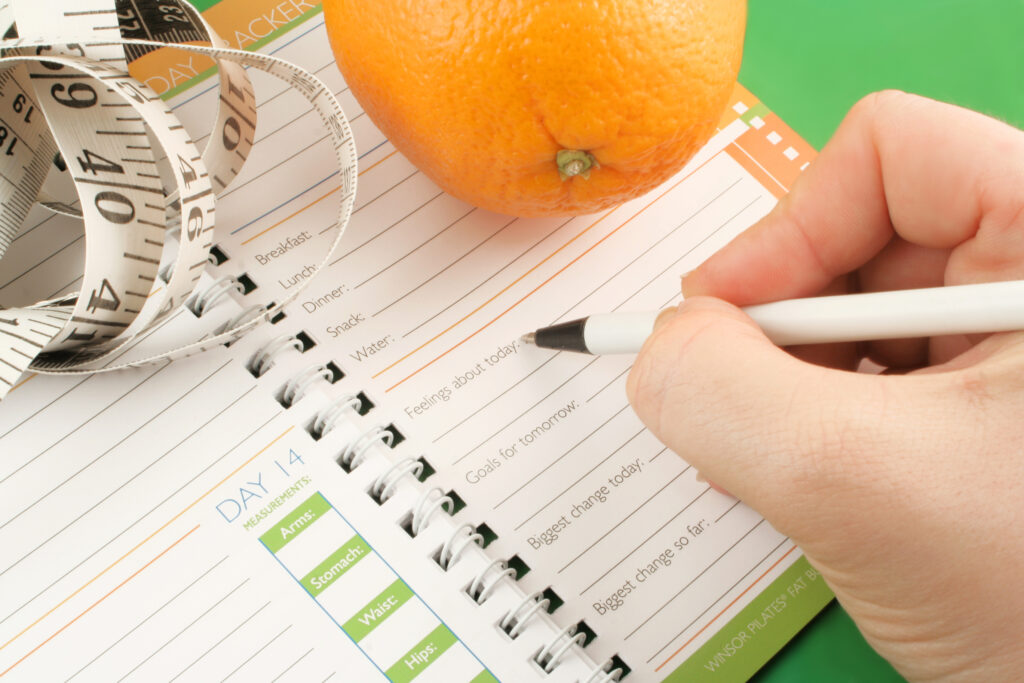 A hand writes in a food and fitness journal beside an orange and a coiled measuring tape, all placed on a green surface.