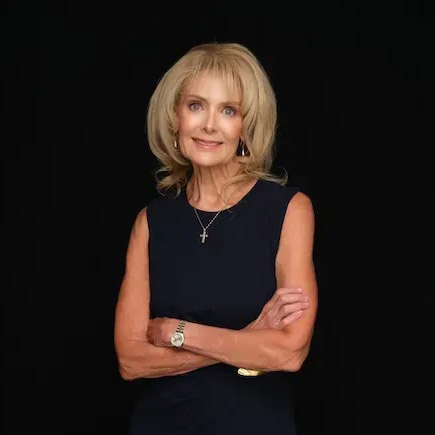 A woman with blonde hair wearing a sleeveless black top, a cross necklace, and a watch stands against a black background with her arms crossed and a slight smile.