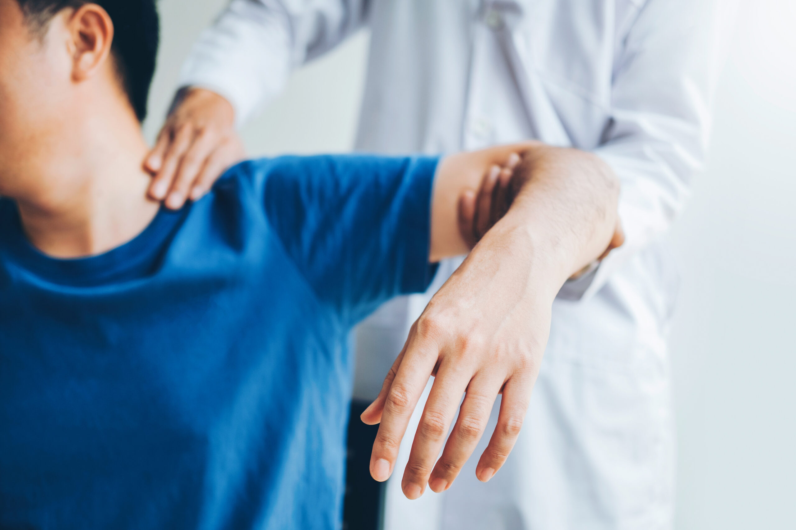 A person in a blue shirt receives shoulder and arm therapy from a healthcare professional wearing a white coat. The professional supports and stretches the person’s arm and shoulder.