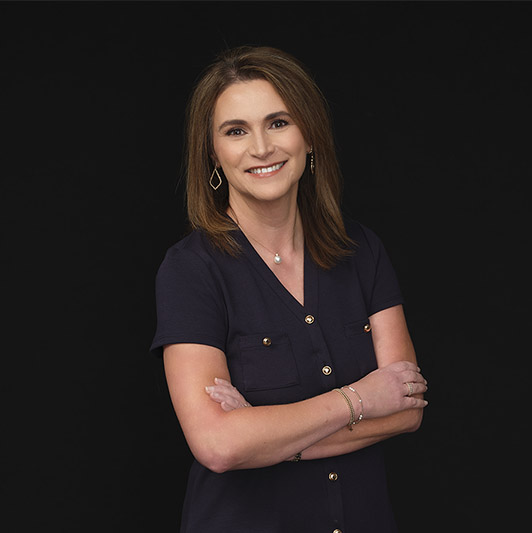 A woman with straight brown hair stands against a black background, smiling with her arms crossed. She wears a navy short-sleeve blouse with gold buttons and simple jewelry.