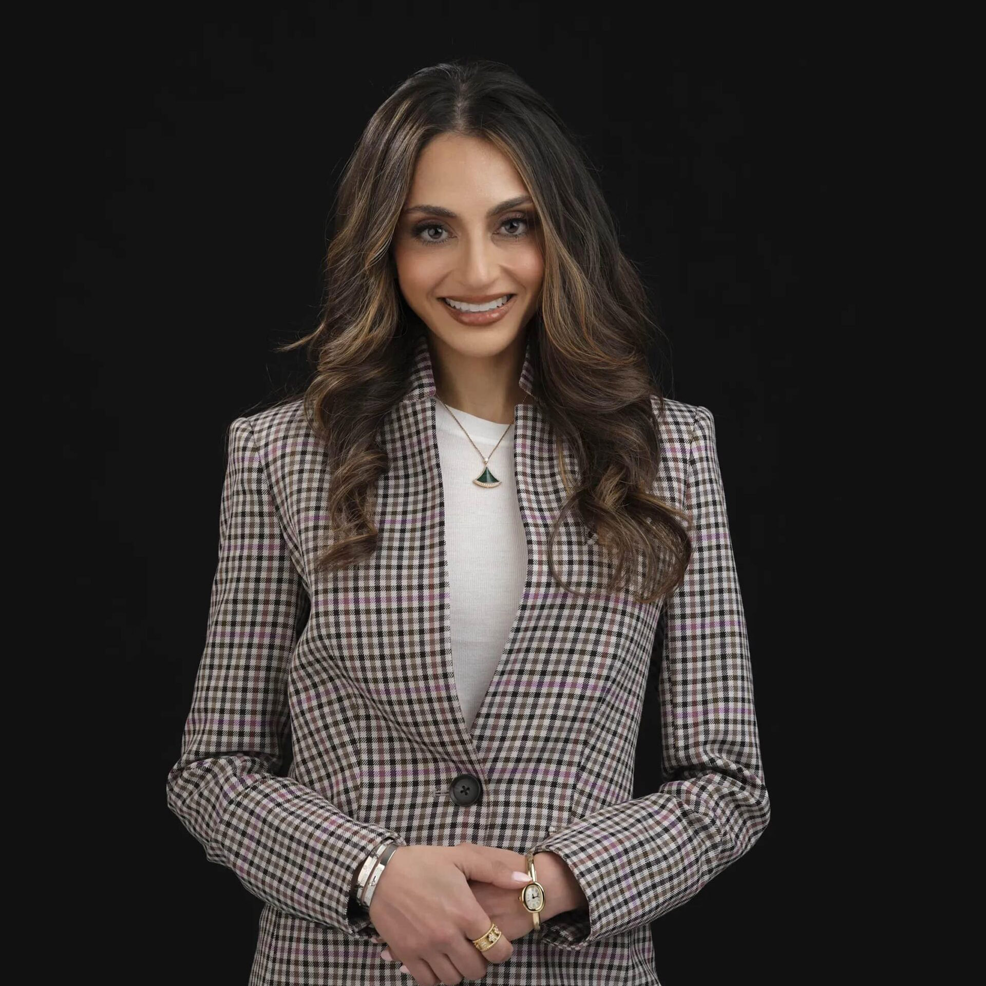 A woman with wavy brown hair wearing a plaid blazer over a white top stands against a black background, smiling with her hands clasped in front. She has jewelry on her wrists and wears a necklace.