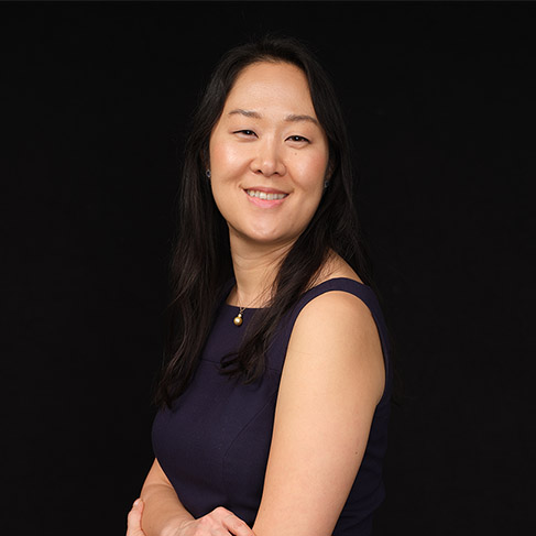 A woman with long dark hair, wearing a sleeveless navy dress, smiles and poses with arms crossed against a plain black background.