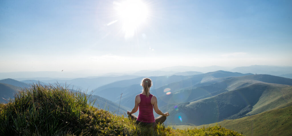 A person sits cross-legged on a grassy hilltop, facing distant mountain ranges under a bright sun and clear blue sky, peacefully meditating in nature.