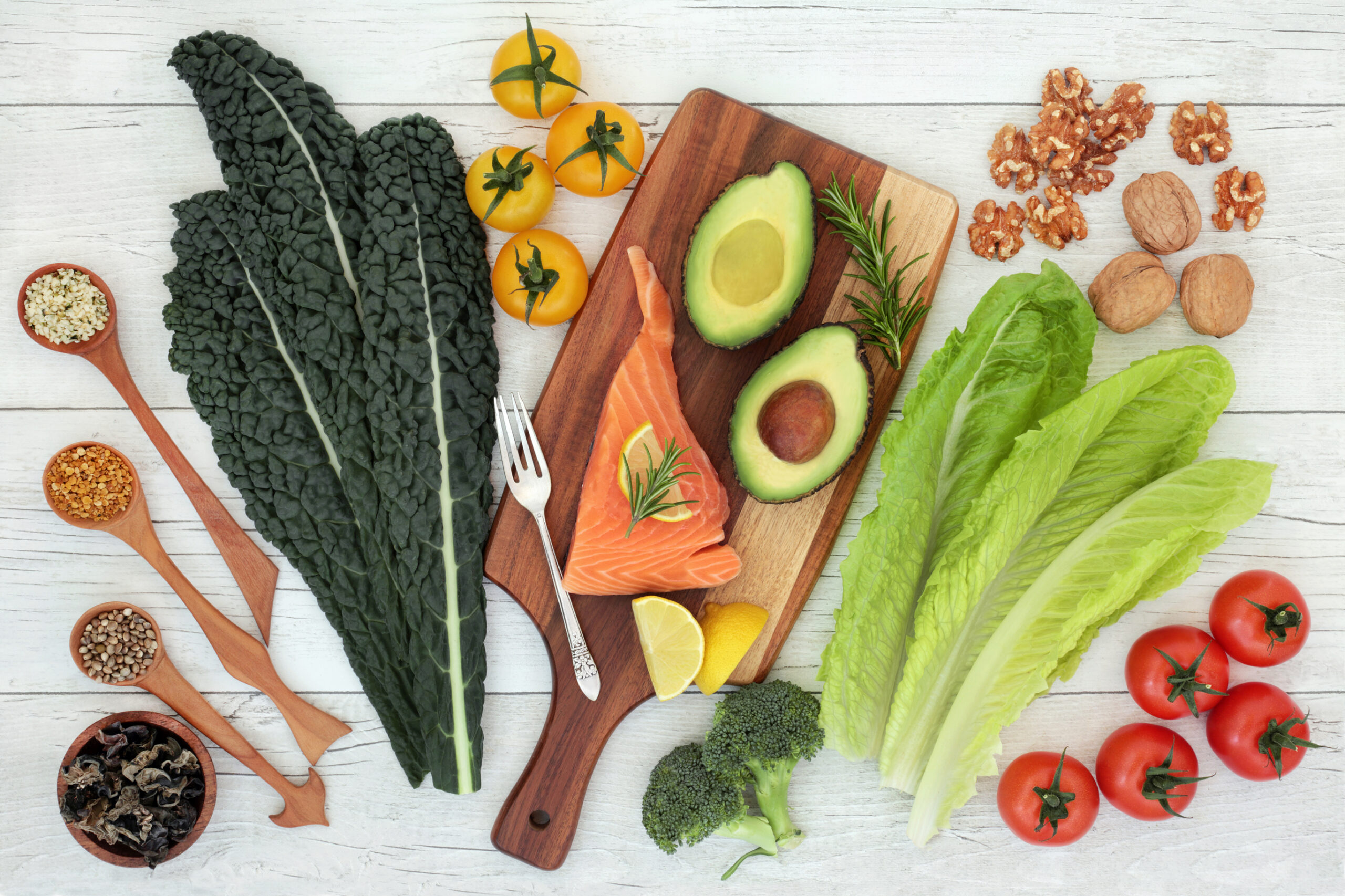 A flat lay of healthy foods including kale, romaine lettuce, cherry tomatoes, broccoli, avocado halves, yellow tomatoes, walnuts, raw salmon, lemon wedges, and wooden spoons with seeds and spices on a white surface.