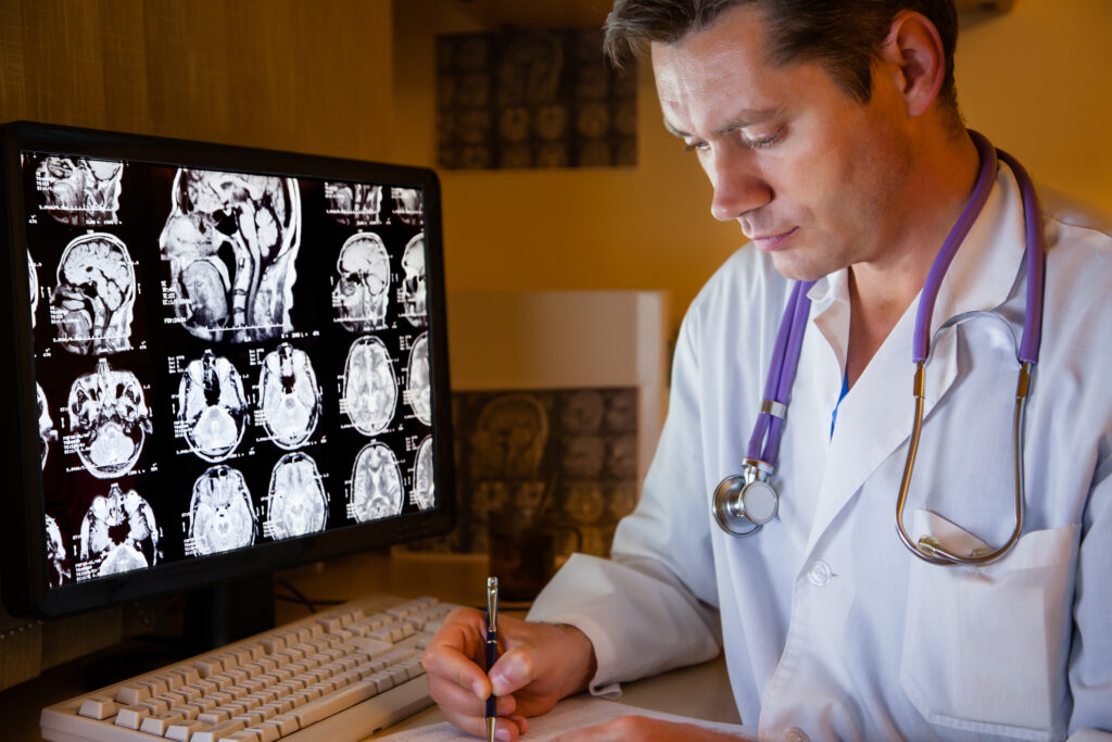 A doctor in a white coat reviews brain scan images displayed on a computer screen while writing notes at a desk, with a stethoscope hanging around his neck.