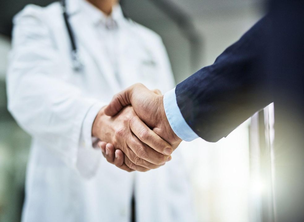 A doctor in a white coat with a stethoscope shakes hands with a person in a suit, symbolizing partnership or agreement in a medical or healthcare setting.