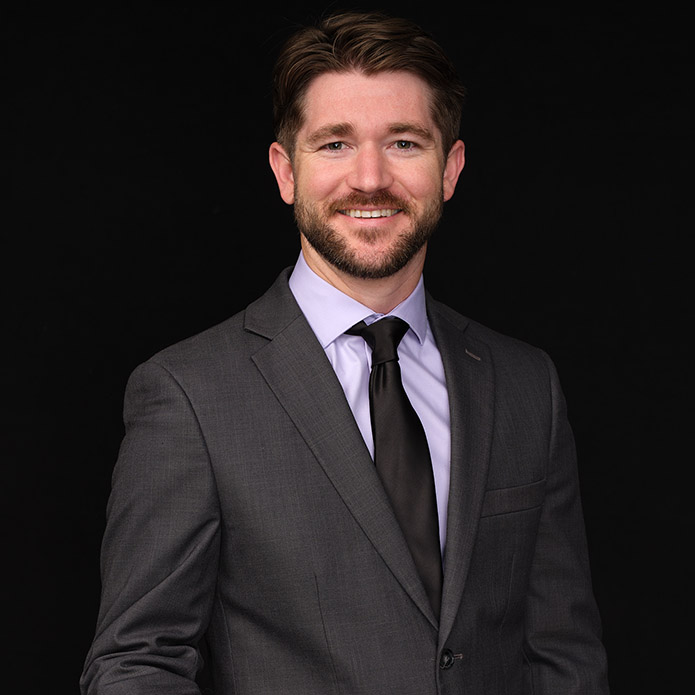 A man with brown hair and a beard wearing a gray suit, light purple shirt, and black tie stands smiling against a plain black background.