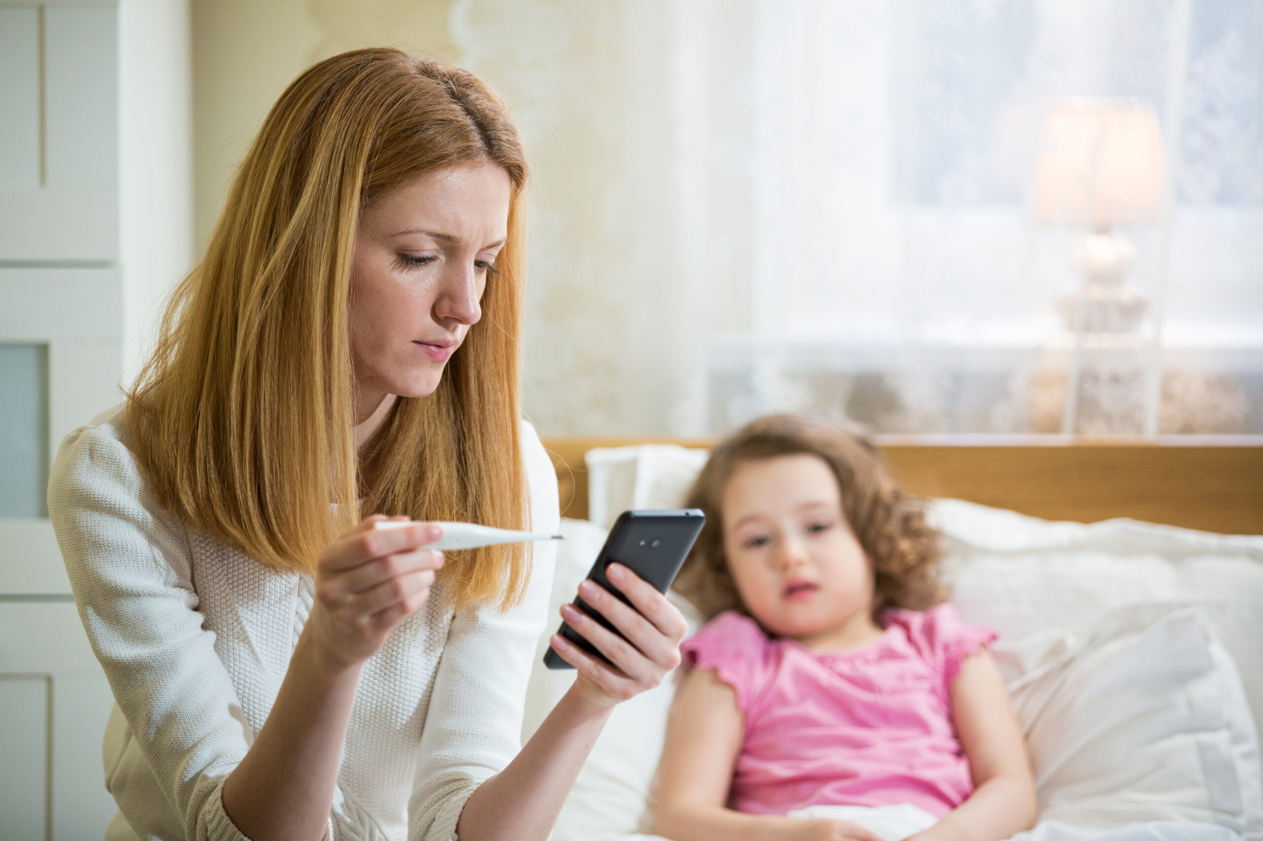 A woman checks her phone while holding a thermometer, sitting beside a young girl in bed who appears unwell, wearing a pink shirt. The room is softly lit with a lamp in the background.