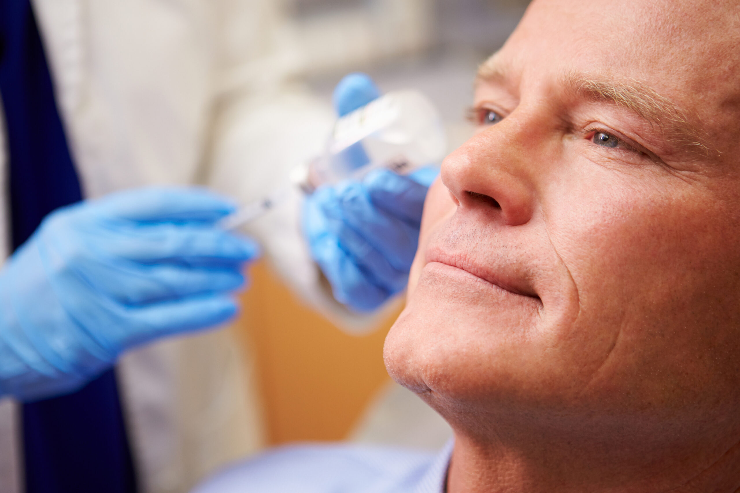 A close-up of a middle-aged man receiving a cosmetic injection in his face from a medical professional wearing blue gloves. The man appears calm and relaxed during the procedure.