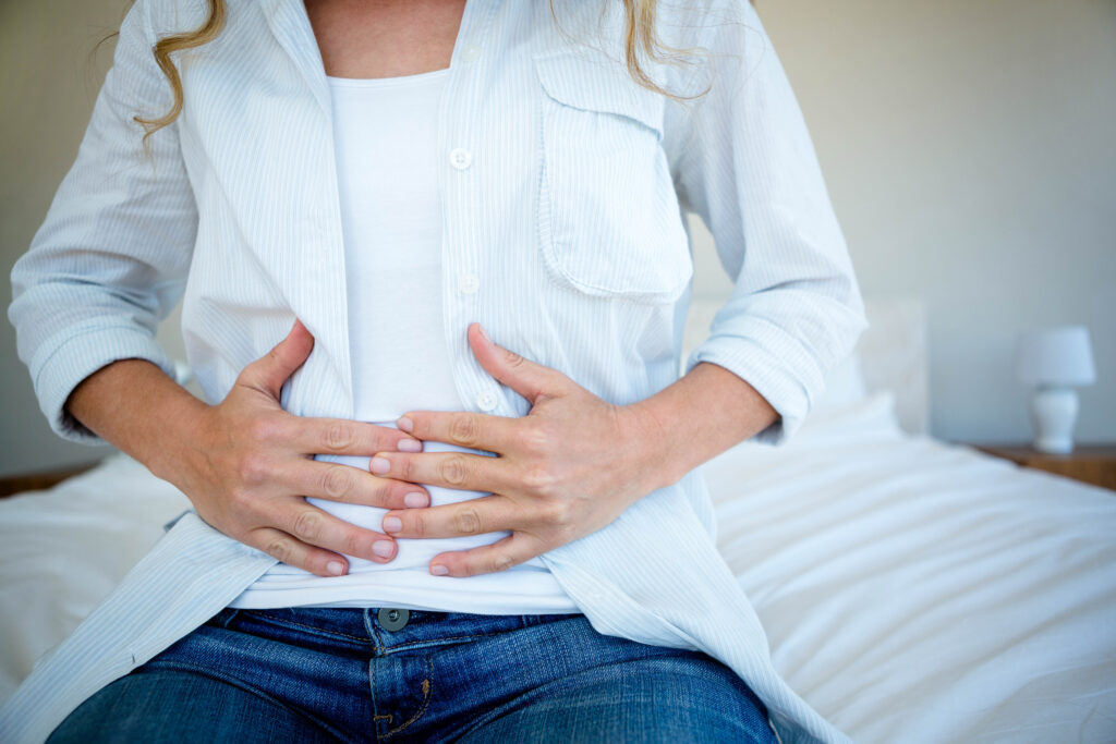A person sitting on a bed, wearing a white shirt and jeans, holding their stomach with both hands, appearing to experience abdominal discomfort or pain.