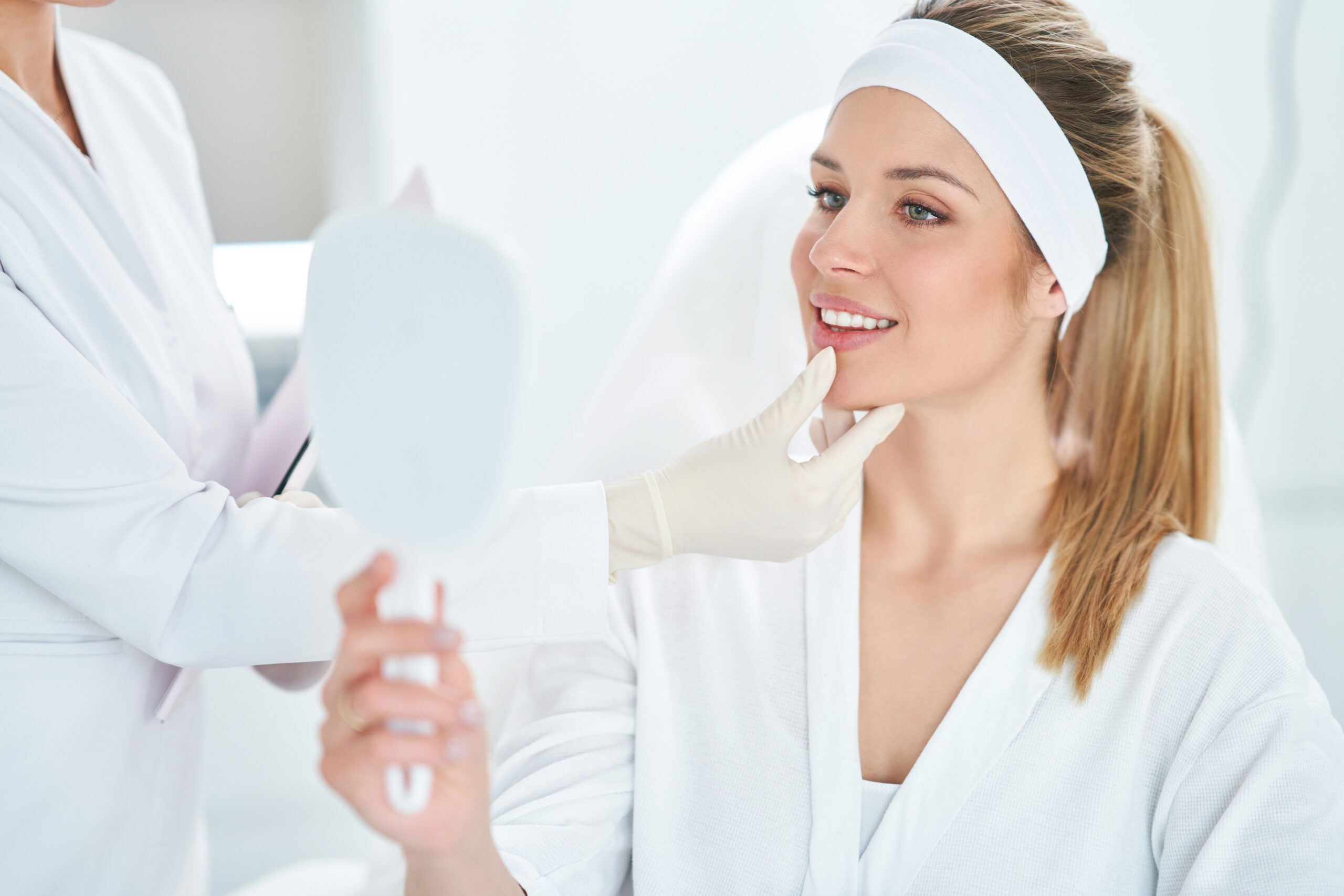 A smiling woman in a white robe and headband looks at herself in a hand mirror while a professional in a white coat and gloves gently touches her chin during a skincare or beauty consultation.