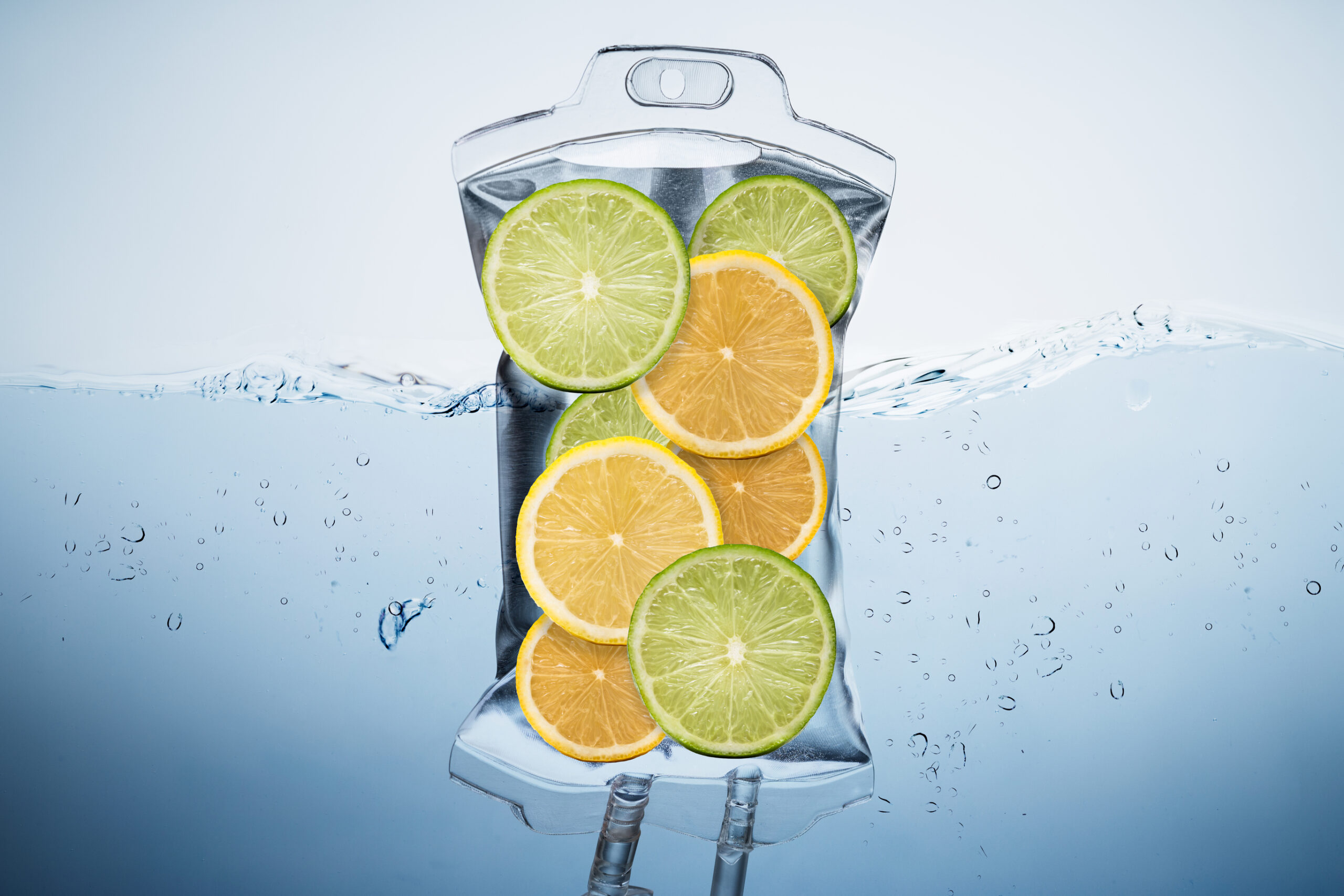 A clear IV bag filled with alternating slices of lime and lemon is submerged halfway in water, with water droplets and splashes visible around the bag. The background is light blue.