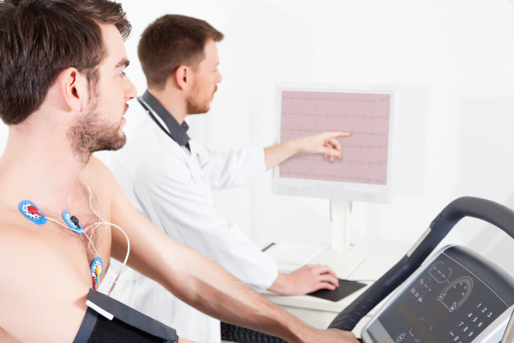 A man with electrodes on his chest uses a treadmill for a cardiac stress test while a doctor in a white coat monitors and points at heart activity data on a computer screen.