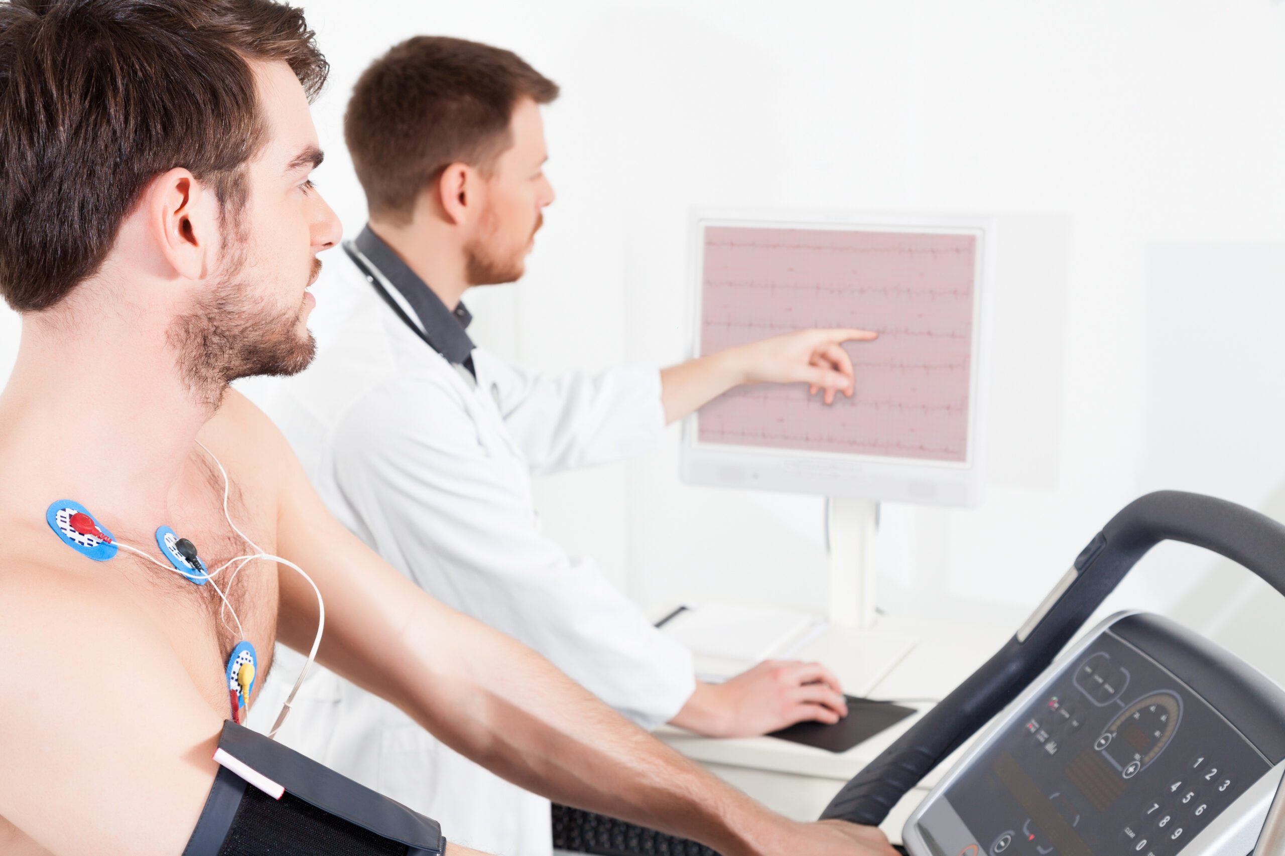 A man with electrodes on his chest uses a treadmill for a cardiac stress test while a doctor in a white coat monitors and points at heart activity data on a computer screen.