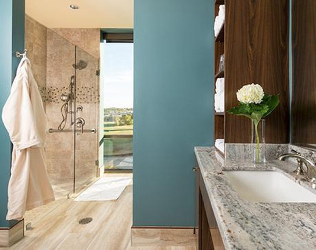 Modern bathroom with blue walls, a granite countertop sink, wooden shelves with white towels, a glass shower with beige tiles, and a white robe hanging on the wall. A vase with a white flower is on the counter.