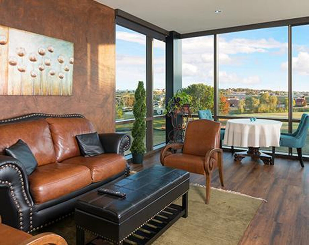 A modern living room with large windows, brown leather furniture, a black ottoman, a round dining table with chairs, potted plants, and a landscape view outside. A painting hangs on the wall above the sofa.