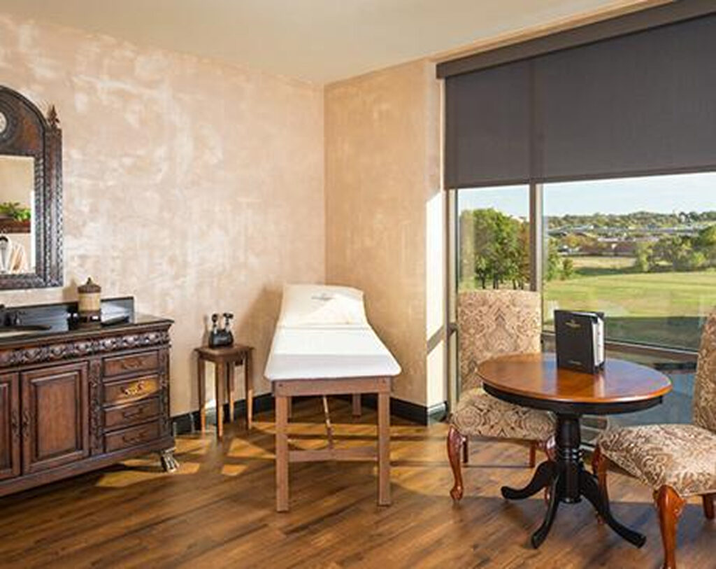 A spa treatment room with a massage table, a wooden dresser with a mirror, two upholstered chairs around a small round table, and large windows overlooking a scenic outdoor view.