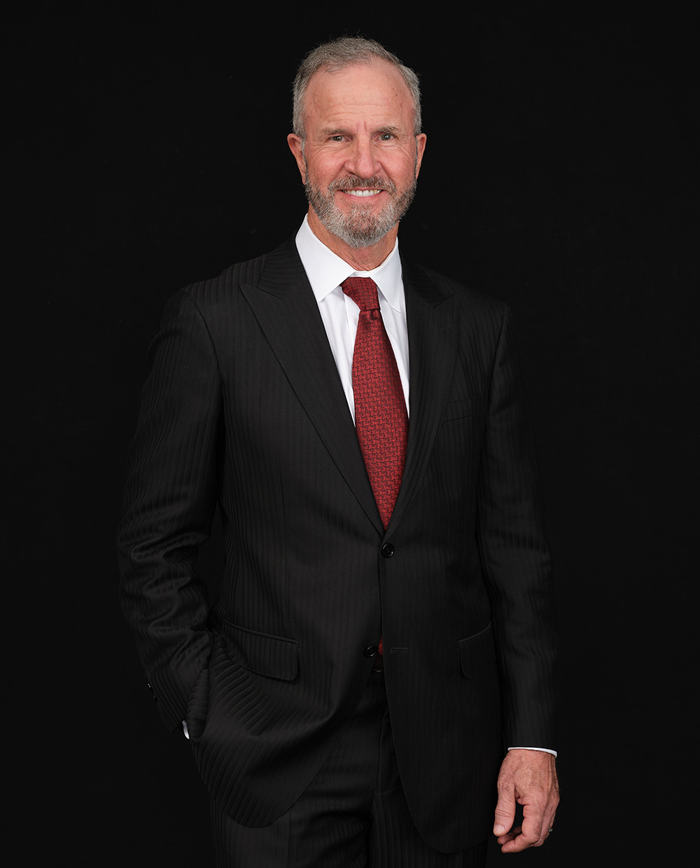 A middle-aged man with gray hair and a beard, wearing a black pinstripe suit, white shirt, and red tie, stands and smiles against a solid black background.