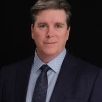 A man with short brown hair wearing a dark suit, blue dress shirt, and patterned tie poses against a plain black background, looking directly at the camera with a slight smile.